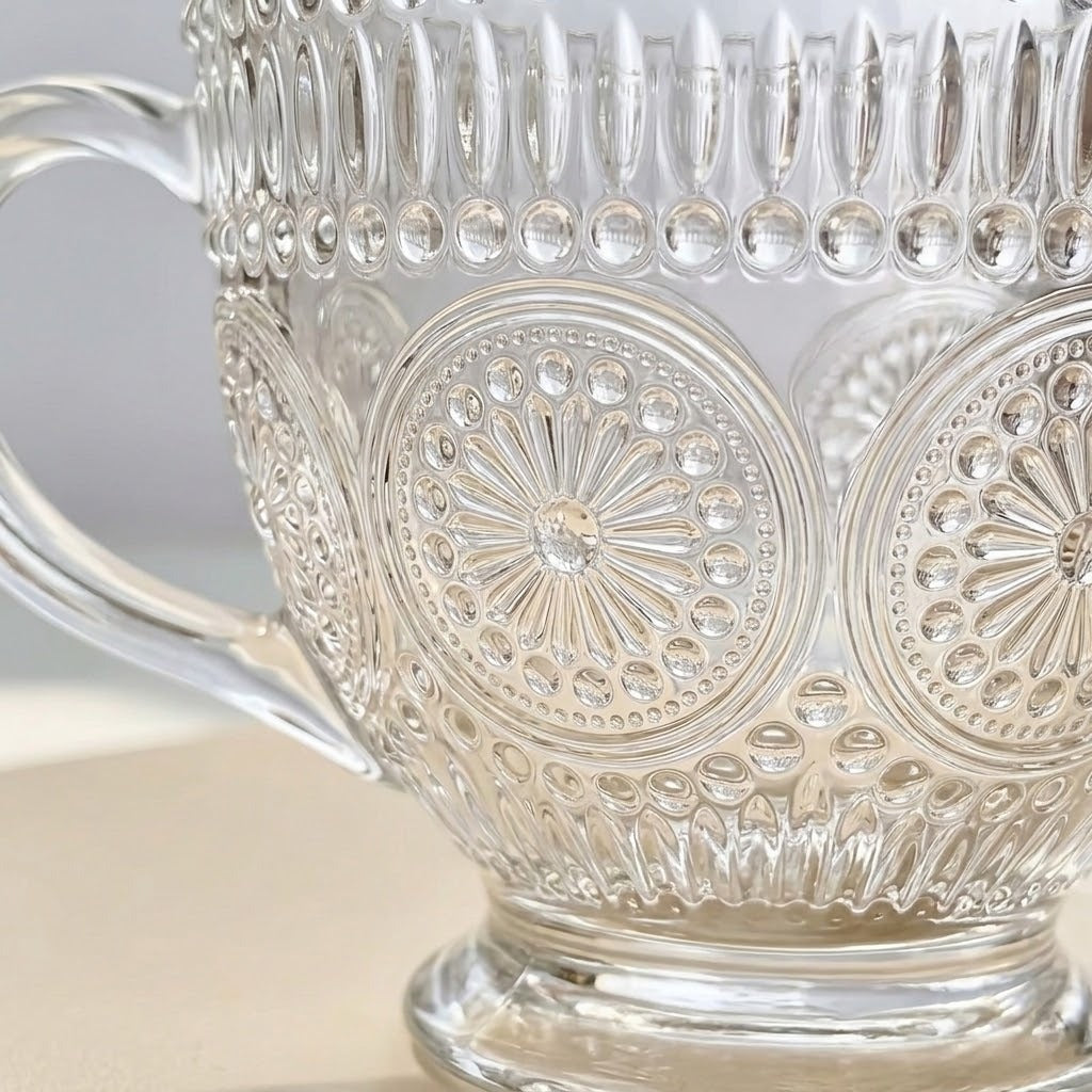 A close-up of a clear glass or acrylic decorative cup with embossed floral and geometric patterns, a side handle, and a pedestal base on a light neutral surface.