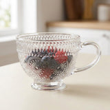 A clear glass teacup with a handle and embossed floral patterns, filled with berries, set on a light beige tabletop with a softly blurred kitchen background.