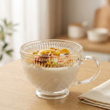 A clear glass mug with a handle and embossed floral pattern, filled with milk and topped with cereal, set on a light wooden table in a softly blurred kitchen setting.