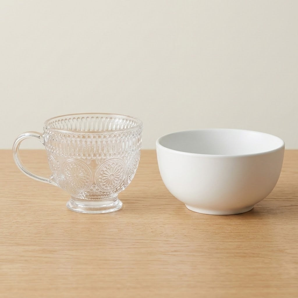A clear glass teacup with embossed floral patterns and a handle, placed next to a white ceramic bowl on a light wood surface against a warm beige background.