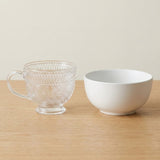 A clear glass teacup with embossed floral patterns and a handle, placed next to a white ceramic bowl on a light wood surface against a warm beige background.