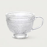 A clear glass teacup with embossed floral and vertical ribbed patterns, set against a solid light gray background.