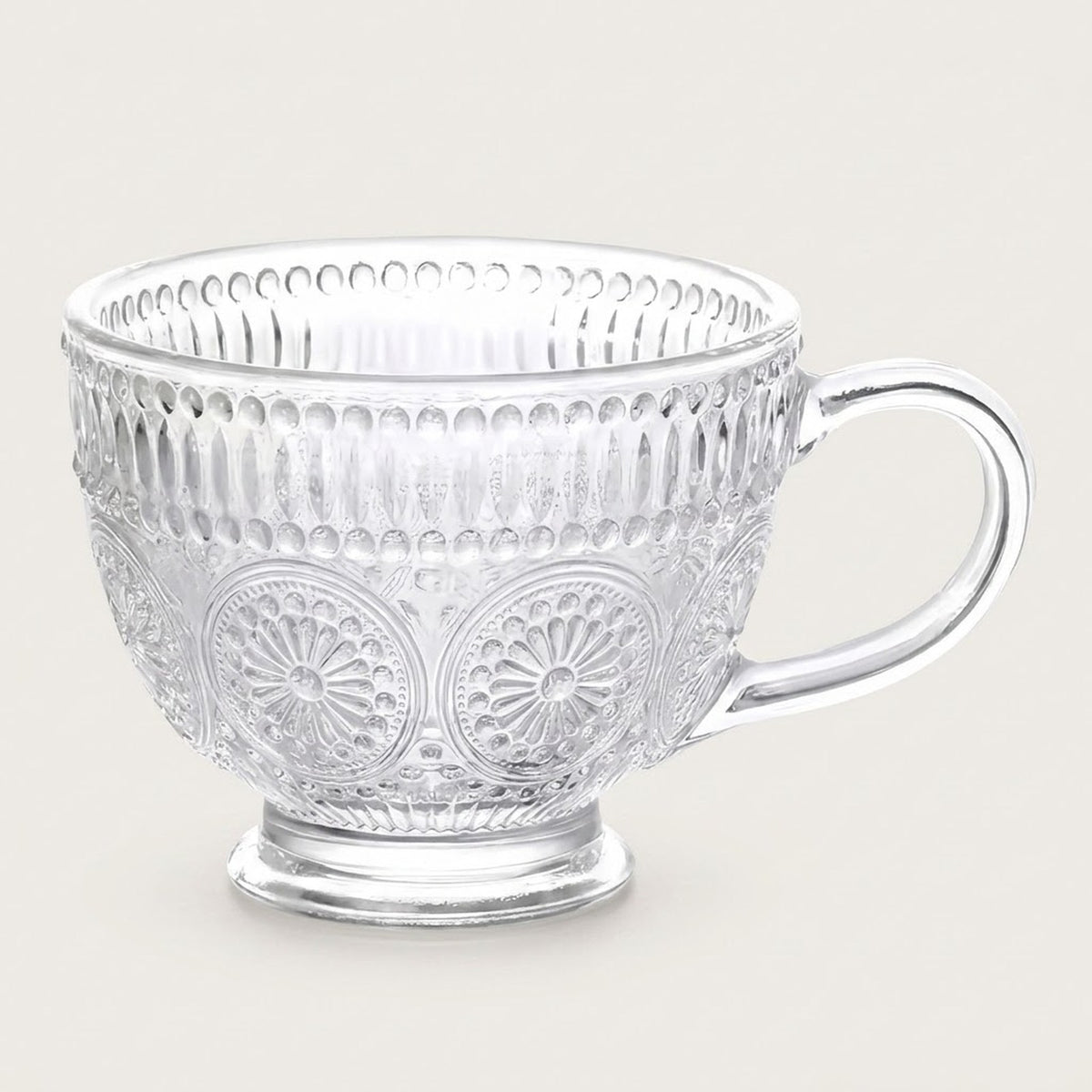 A clear glass teacup with embossed floral and geometric patterns, set against a solid light beige background.