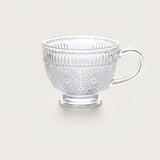 A clear glass teacup with embossed floral and vertical ribbed patterns, set against a solid light beige background.