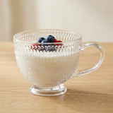 A clear glass teacup with a decorative beaded rim and embossed floral pattern, filled with white yogurt and topped with blueberries and a strawberry, set on a light wood surface.