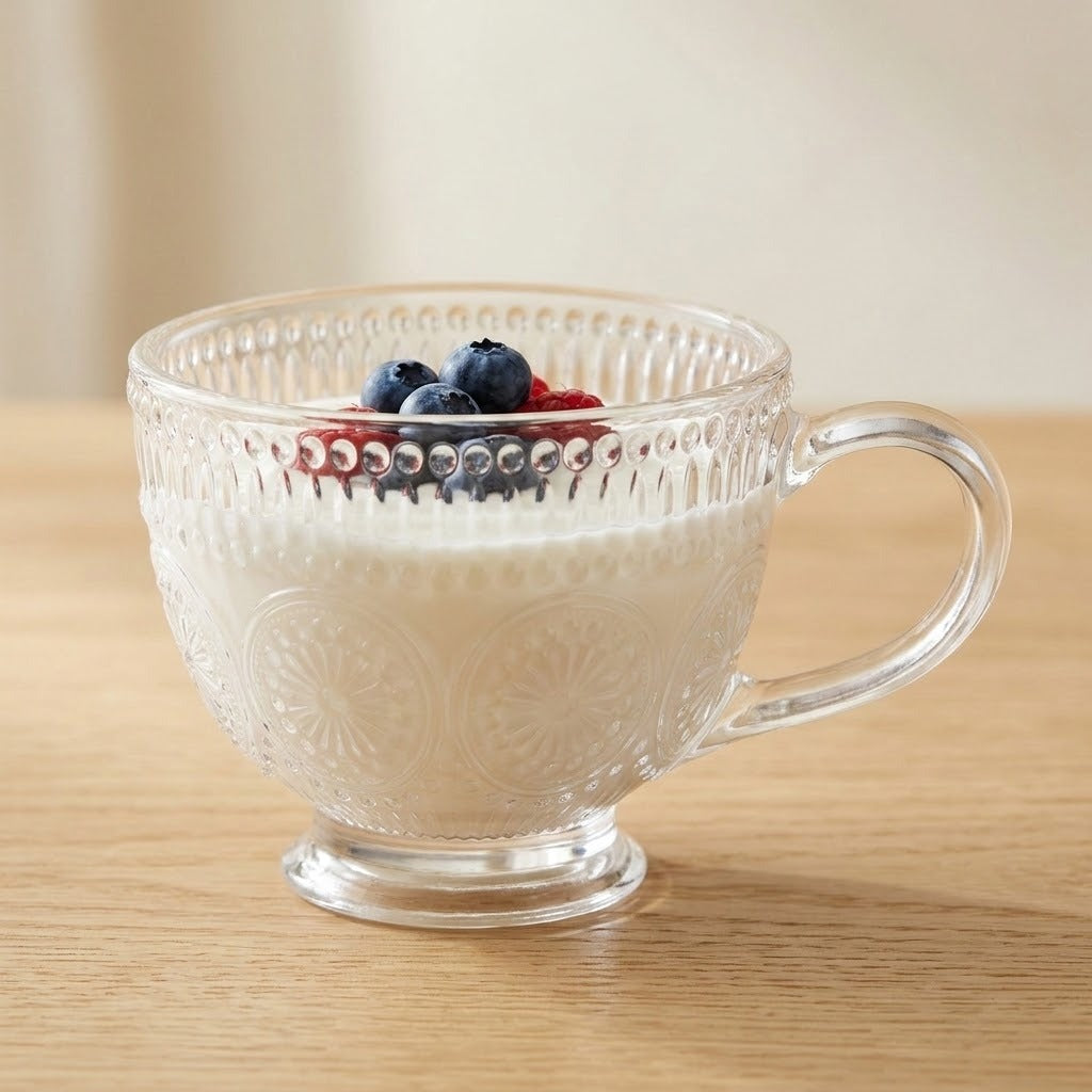 A clear glass teacup with a decorative beaded rim and embossed floral pattern, filled with white yogurt and topped with blueberries and a strawberry, set on a light wood surface.