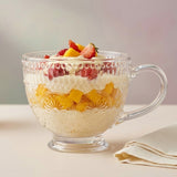 A clear glass dessert cup with a handle, filled with layered cream, fruit pieces, and strawberries, set on a light surface with a folded napkin nearby.
