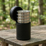 A black textured cylindrical lunch bag with four nested stainless steel compartments, shown open on a rustic wooden picnic table with a blurred forest background.
