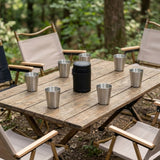 A rustic wooden folding table set in a forest with canvas camping chairs around it. On the table are several stainless steel cups and a black cylindrical container.