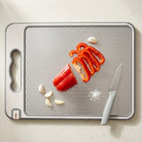 A gray textured rectangular cutting board with a built-in handle, shown on a light beige countertop. On the board are sliced red bell peppers, garlic cloves, a white-handled knife, and a small pile of salt.