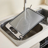 A rectangular stainless steel cutting board with a black handle and rim, shown in a sink under running water with a dishwasher visible in the background.