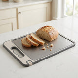 A rectangular gray and silver cutting board with a handle, shown on a white kitchen countertop with sliced bread and a loaf placed on top.