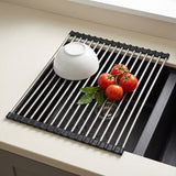 A black and stainless steel collapsible sink drying rack placed on a kitchen sink, with a white bowl and red tomatoes on top.