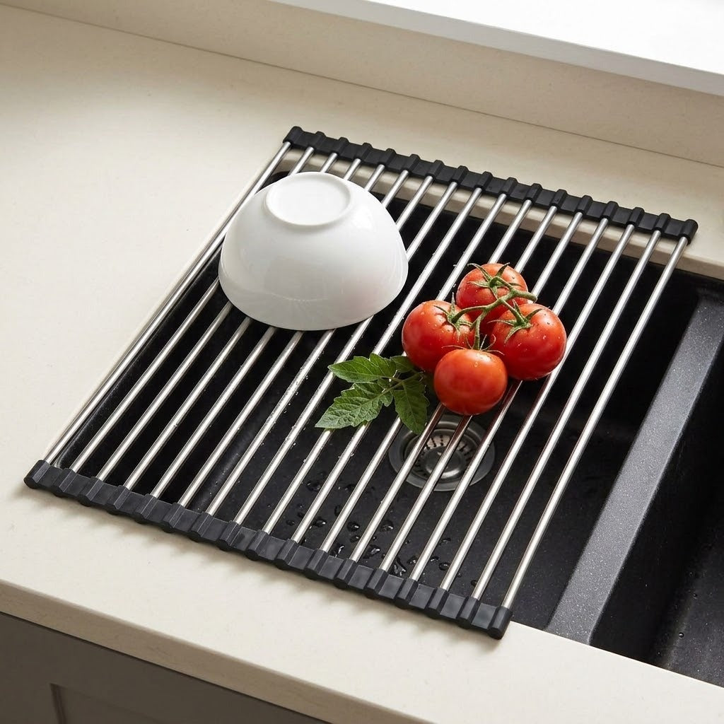 A black and stainless steel collapsible sink drying rack placed on a kitchen sink, with a white bowl and red tomatoes on top.