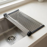 A stainless steel collapsible sink drain guard with black plastic end caps, shown draped over the edge of a stainless steel kitchen sink on a white countertop.
