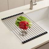 A stainless steel sink drain guard with black rubber end caps, shown in a white kitchen sink with lettuce and a red bell pepper on top.
