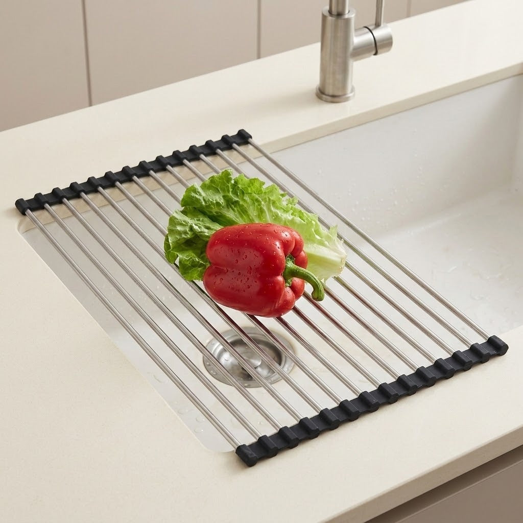 A stainless steel sink drain guard with black rubber end caps, shown in a white kitchen sink with lettuce and a red bell pepper on top.