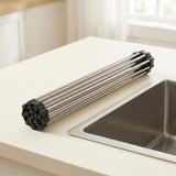 A stainless steel and black plastic kitchen utensil with multiple parallel rods, resting on a white countertop next to a stainless steel sink in a bright kitchen.