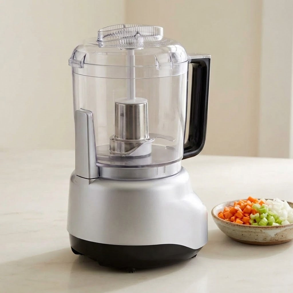 A countertop food processor with a clear plastic bowl, silver base, and black handle and bottom trim, shown on a light beige surface with a small bowl of chopped vegetables beside it.