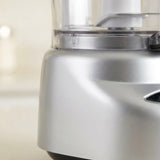 Close-up of a silver countertop food processor with a clear bowl and metallic base, shown against a softly blurred light background.