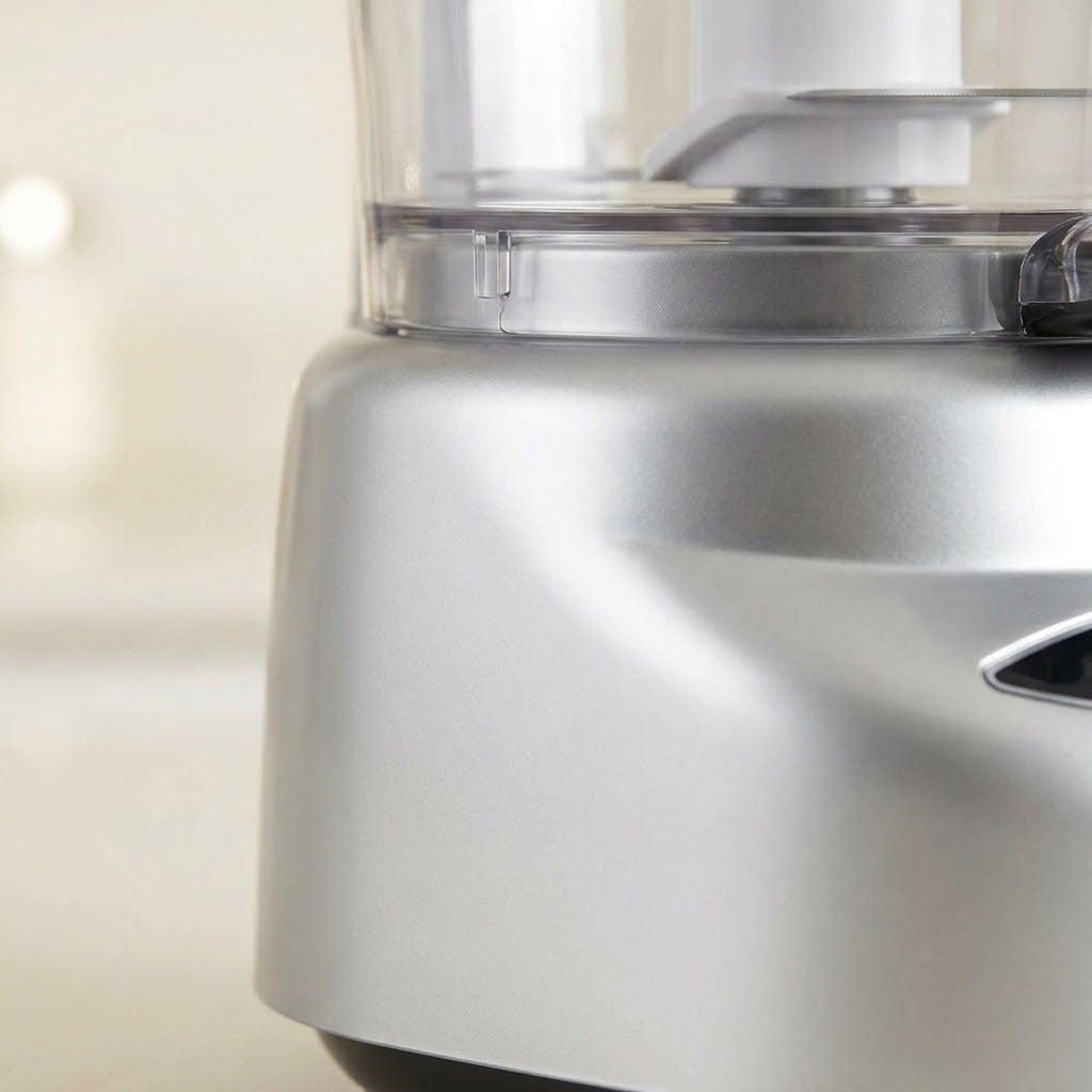 Close-up of a silver countertop food processor with a clear bowl and metallic base, shown against a softly blurred light background.