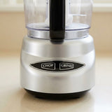 A close-up of a countertop food processor with a brushed silver metal base, black buttons labeled CHOP and GRIND, and a clear plastic bowl with a black handle.