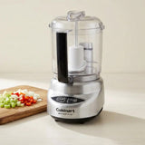 Cuisinart Mini Prep Plus food processor with a clear container, silver base, and black handle, shown on a light countertop with chopped vegetables on a cutting board.