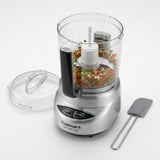 Cuisinart food processor with a clear bowl filled with chopped vegetables, shown with the clear lid off and a gray spatula on a white background.
