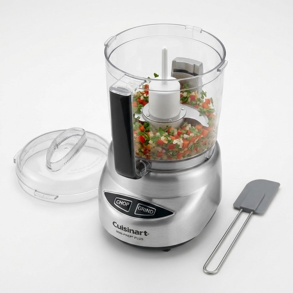 Cuisinart food processor with a clear bowl filled with chopped vegetables, shown with the clear lid off and a gray spatula on a white background.