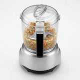 A countertop food processor with a clear plastic bowl filled with chopped vegetables, a silver metal base, and a black handle, shown on a clean white background.