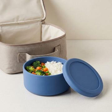 A blue silicone bento container with a matching lid, shown open with white rice and broccoli on top, with an open beige insulated lunch bag in the background on a light surface.