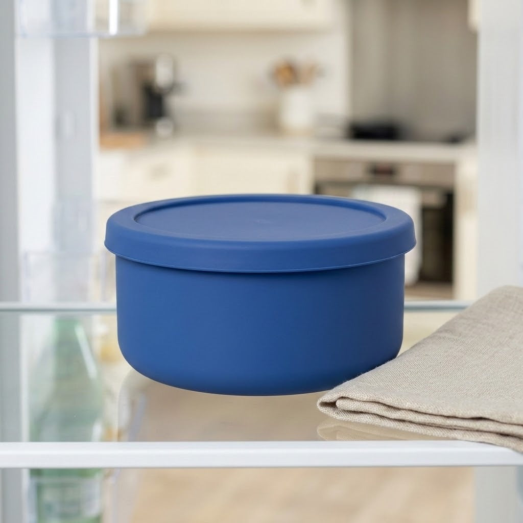 A blue round silicone storage container with a matching lid, shown on a glass shelf next to a folded beige cloth, with a blurred kitchen background.