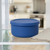 A blue round silicone storage container with a matching lid, shown on a glass shelf next to a folded beige cloth, with a blurred kitchen background.