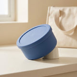 A blue round lidded container with a matching lid, photographed on a light beige surface in a softly lit interior setting with a cream tote bag blurred in the background.