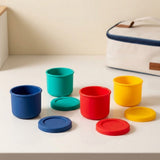 Six small cylindrical storage containers in blue, teal, red, and yellow, shown with matching lids on a light tabletop. A cream-colored zippered bag with blue trim is partially visible in the background.
