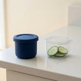 A dark blue silicone food storage container with a matching lid sits on a light speckled countertop next to a clear plastic container holding cucumber slices.