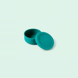 A small round teal container with its matching lid leaning against it, photographed on a clean pale mint background.