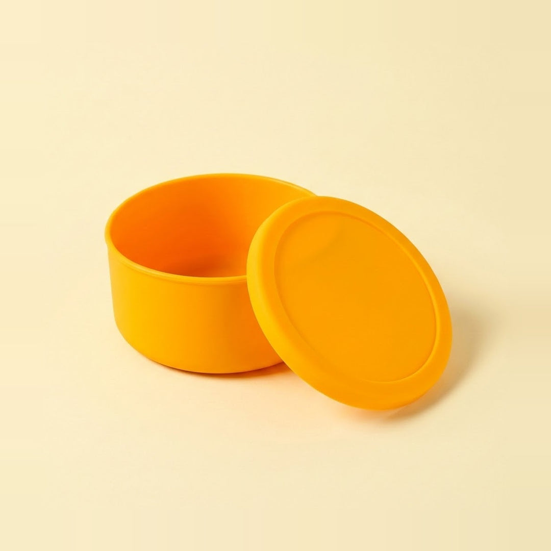 A bright yellow-orange silicone container with its matching lid leaning against it, set against a solid cream-colored background.