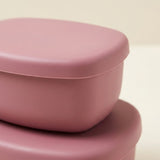 A close-up of a dusty pink plastic container with a matching lid, stacked on top of another container against a light cream background.
