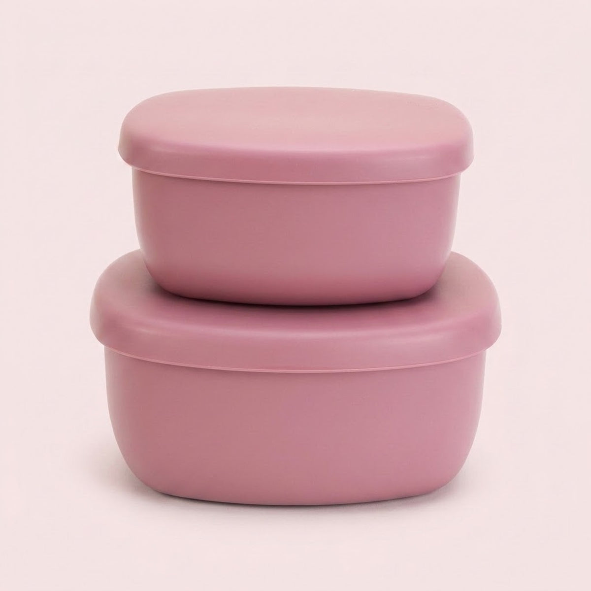 Two stacked dusty pink silicone food storage containers with matching lids, set against a solid pale pink background.