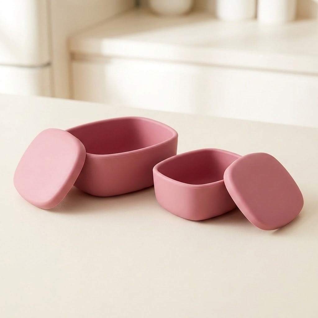 Two small pink silicone containers with matching lids, shown open on a light cream tabletop in a softly blurred indoor setting.