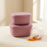 Two stacked dusty pink plastic food storage containers on a light cream surface, with one container tipped over spilling a small amount of orange food. A soft-focus indoor background is visible.