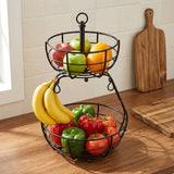 A two-tier black wire fruit basket filled with fresh fruit, including bananas, apples, oranges, and tomatoes, set on a wooden kitchen counter with a cutting board in the background.
