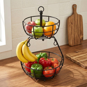 A two-tier black wire fruit basket filled with fresh fruit, including bananas, apples, oranges, and tomatoes, set on a wooden kitchen counter with a cutting board in the background.