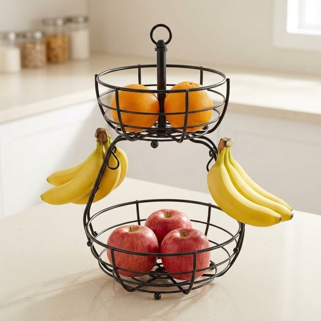 A two-tier black wire fruit basket filled with oranges on the top tier and red apples and bananas on the lower tier, set on a kitchen counter.