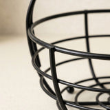 Close-up of a black metal wire basket or planter with a rounded oval shape, photographed against a light beige background.