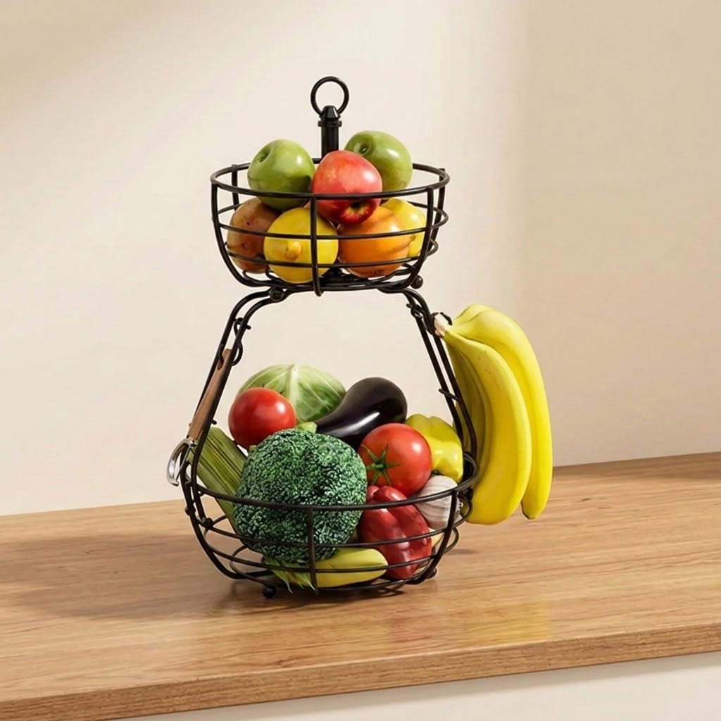 A two-tier black wire fruit basket filled with fresh fruits including bananas, broccoli, tomatoes, and apples, displayed on a wooden countertop against a beige wall.