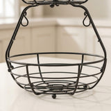 Black metal wire basket with a top tier and hanging loops, shown on a light countertop with a blurred kitchen background.