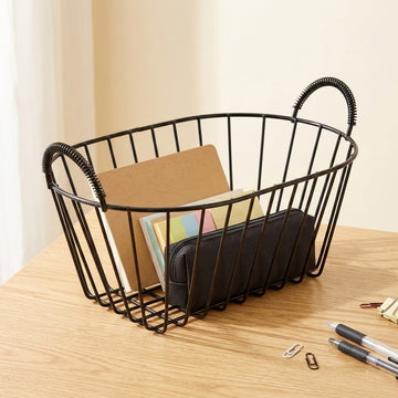 A black wire basket organizer with two loop handles sits on a light wood desk, holding notebooks and a black pouch. Pens and paper clips are visible on the desk surface in the foreground, with a soft beige background.