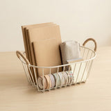 A white wire basket with rope handles sits on a light wood table, holding kraft paper folders, a beige fabric pouch, and rolls of washi tape.
